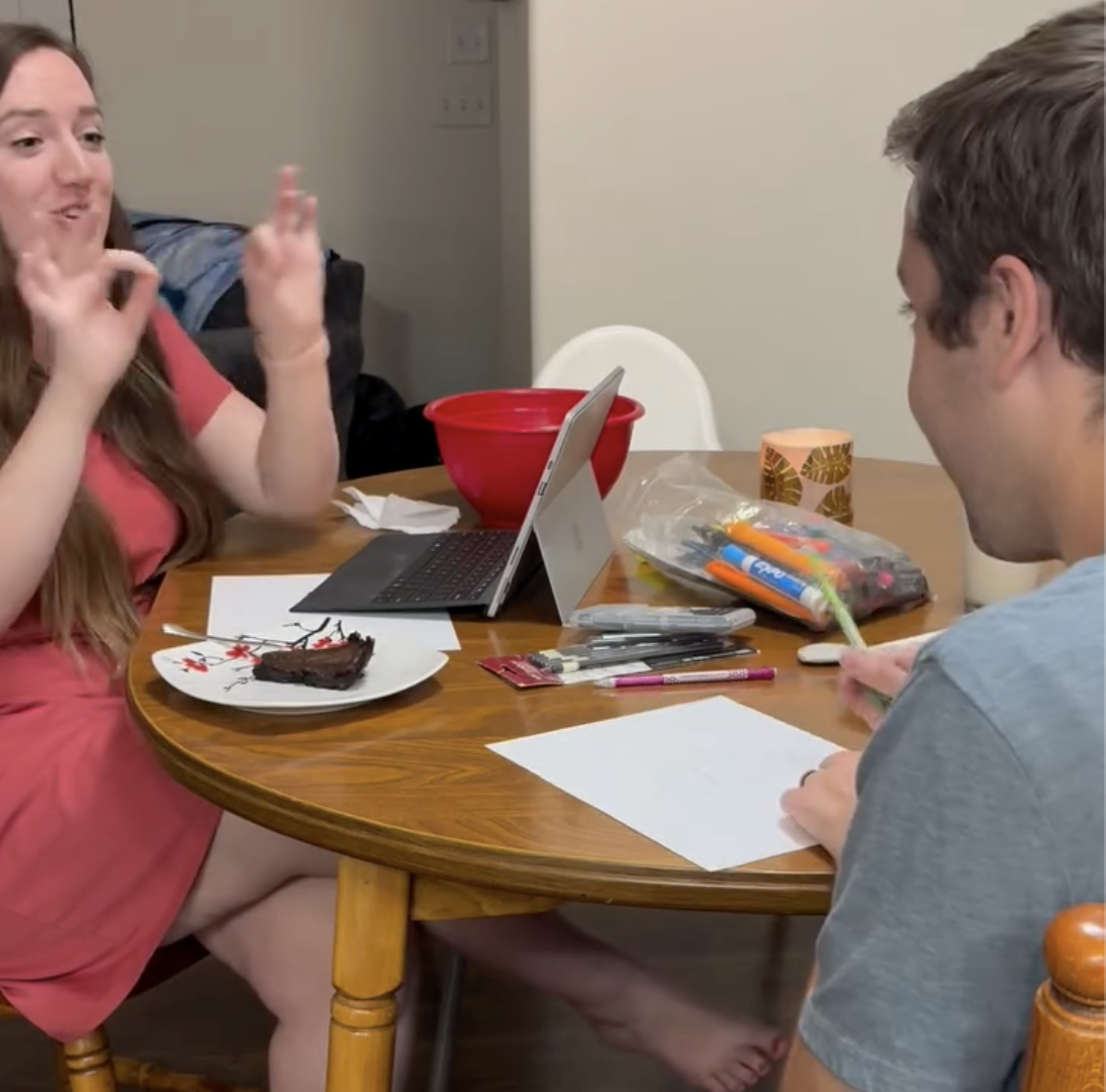 Wife describing a drawing tutorial out loud while her husband draws it blind during an at-home drawing date night