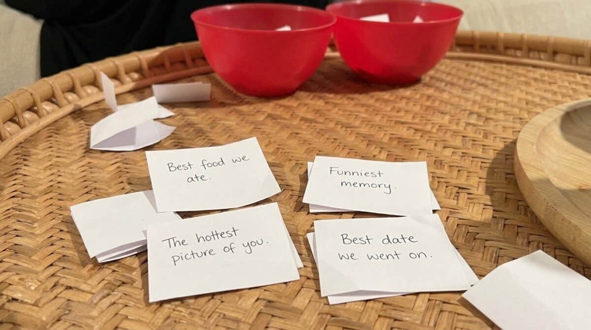 Two bowls in the background. A close-up of slips of paper that say prompts like: best food we ate, funniest memory, the hottest picture of you, best date we went on.